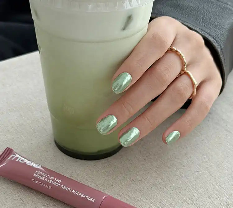 Parlak açık yeşil ojeli bir elin, ortasında bir matcha latte tutarken, ön planda bir pembe dudak parlatıcısı tüpü.