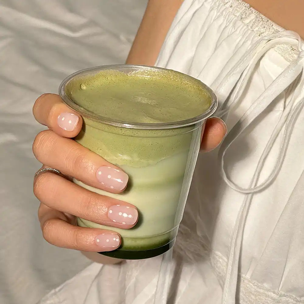 Beyaz puantiyeli beyaz ojeli, köpüklü üst kısmı ve katmanlı matcha latte içeren şeffaf plastik bir bardak tutan el.